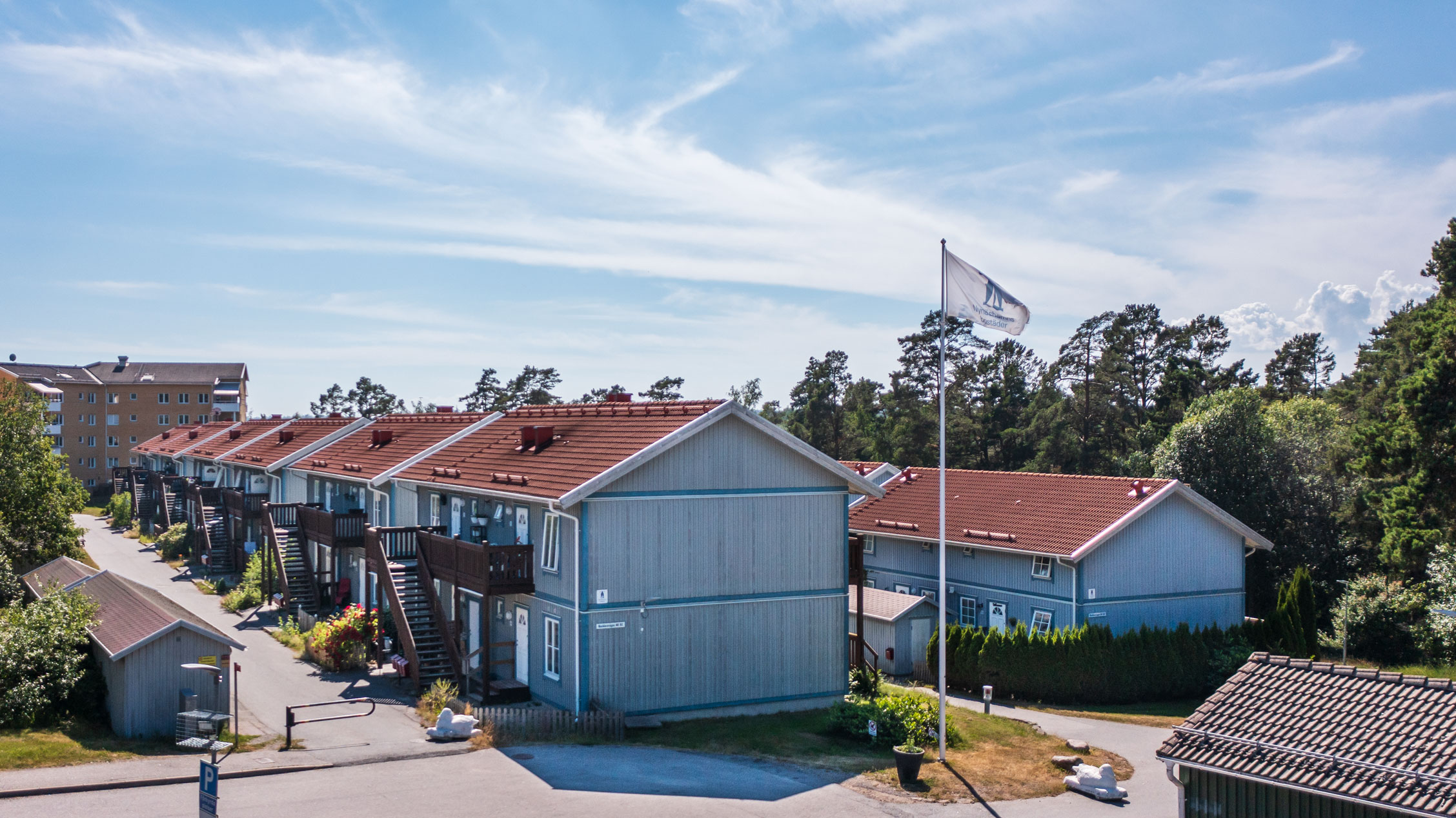 Rad av gråa radhus med röda tak längs Backluravägen 30 till 112 i Nynäshamn. Husen har utvändiga trappor och små trädgårdar. En flagga med Nynäshamnsbostäders logo vajar i förgrunden och flerfamiljshus syns i bakgrunden.