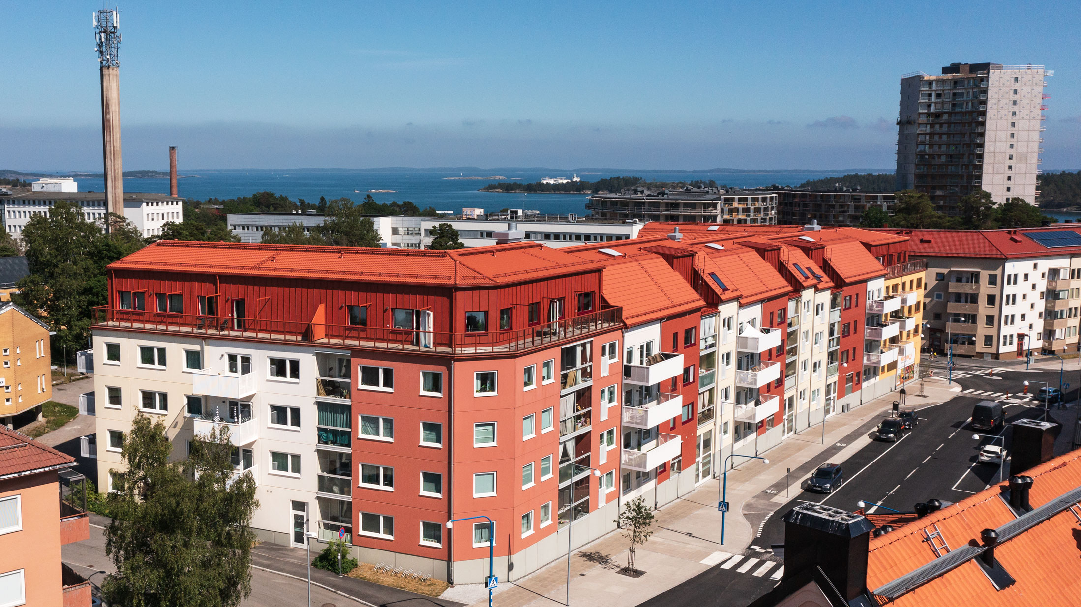 Moderna flerbostadshus med färgglada fasader på Bryggargatan 2 och Nynäsvägen 14 i Nynäshamn. Byggnaderna ligger nära havet med utsikt över omgivande landskap och havet i bakgrunden. Gatan är livlig med bilar och tydliga gångvägar.