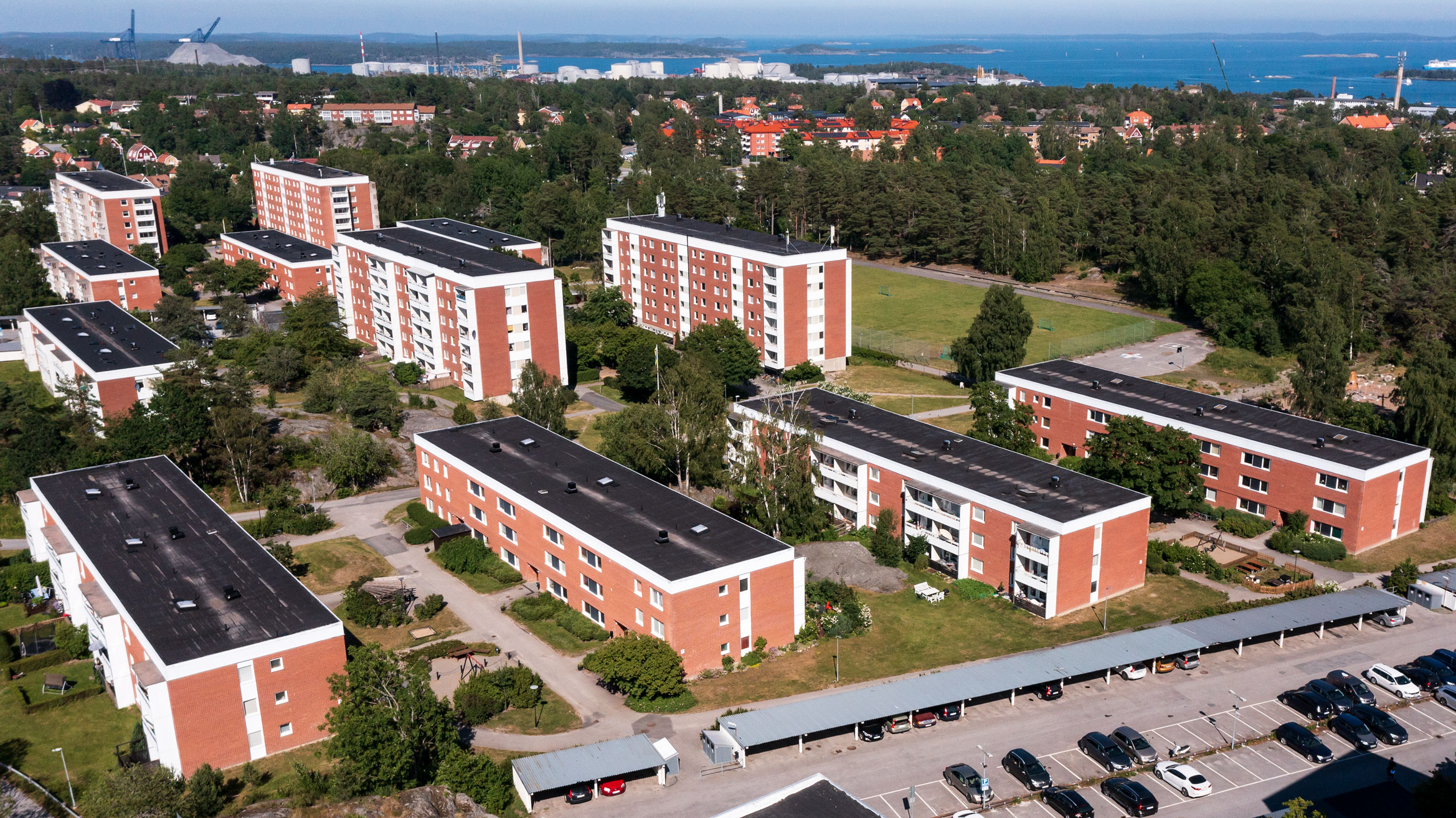 Flera flerbostadshus på Backluravägen 3 till 25 i Nynäshamn, omgivna av grönska med utsikt över havet i bakgrunden. Parkeringsplatser och lekplats synliga i förgrunden.