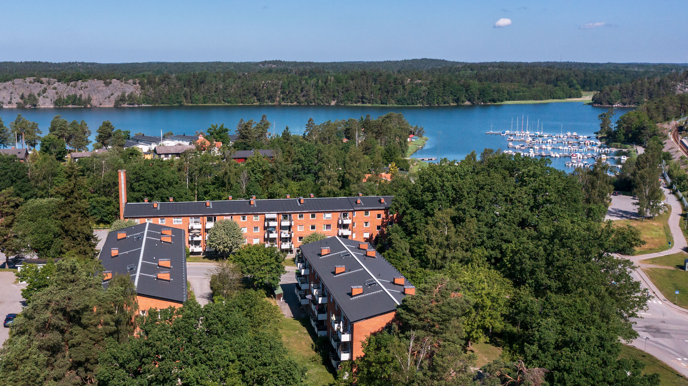 Flygbild över hyreslägenheter på Estövägen 1, 2 och 3 i Nynäshamn, med röda tegelbyggnader omgivna av träd och grönska, nära vattnet och småbåtshamnen. Perfekta hyreslägenheter i en naturskön miljö.