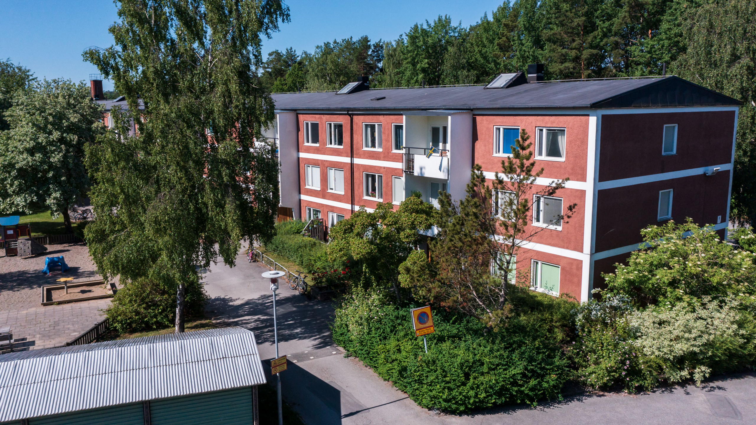 Närbild av hyreslägenheter på Estövägen 7 och 9 i Nynäshamn, med röda fasader och balkonger, omgivna av träd och grönska. Perfekta hyreslägenheter nära en lekplats och natur.