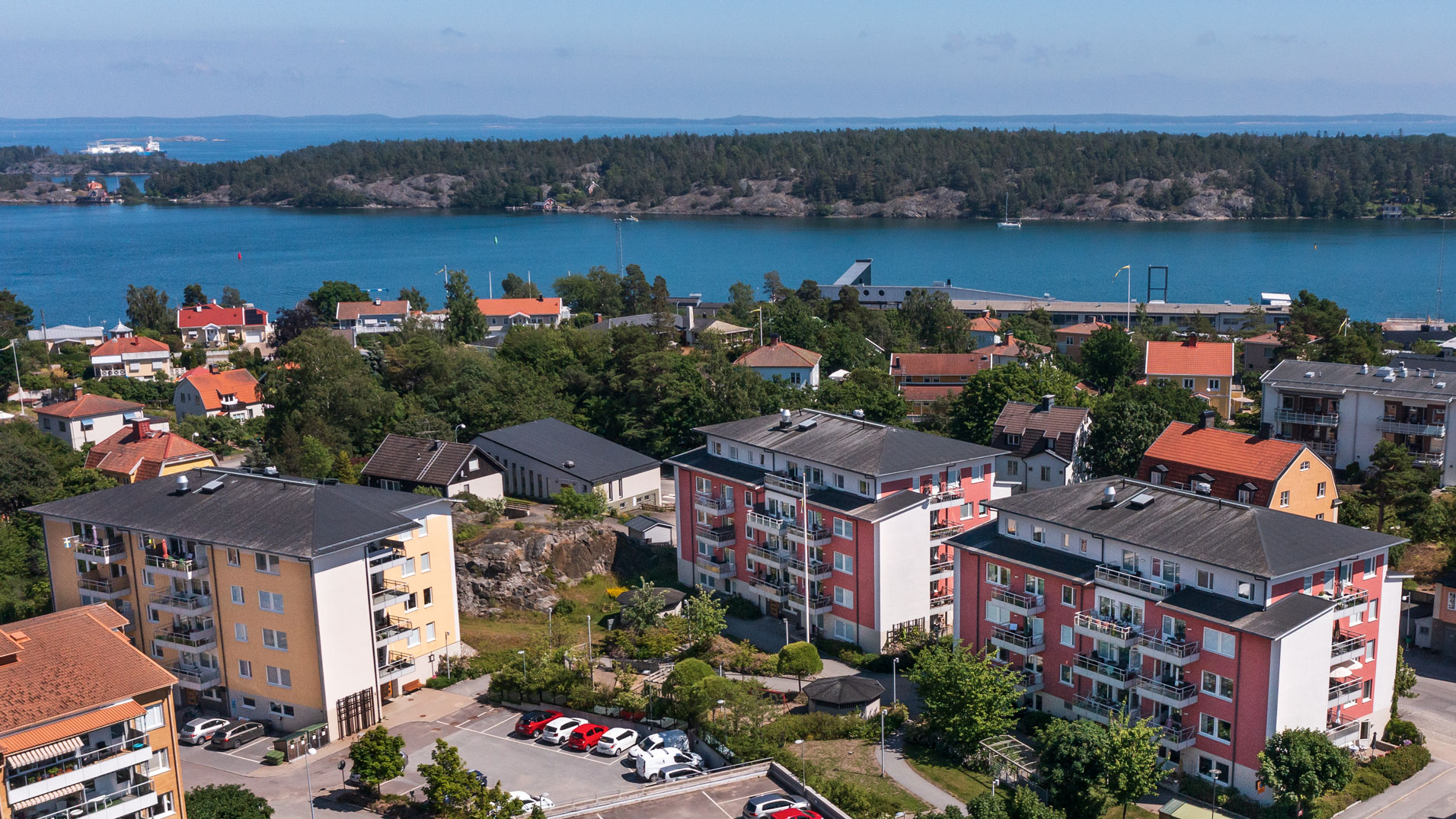 Flygbild över hyreslägenheter på Åkargränd 4, Fredsgatan 19 och 21 i Nynäshamn, med färgglada byggnader och balkonger, omgivna av grönska och med närhet till vatten. Perfekta hyreslägenheter med utsikt över skärgården.