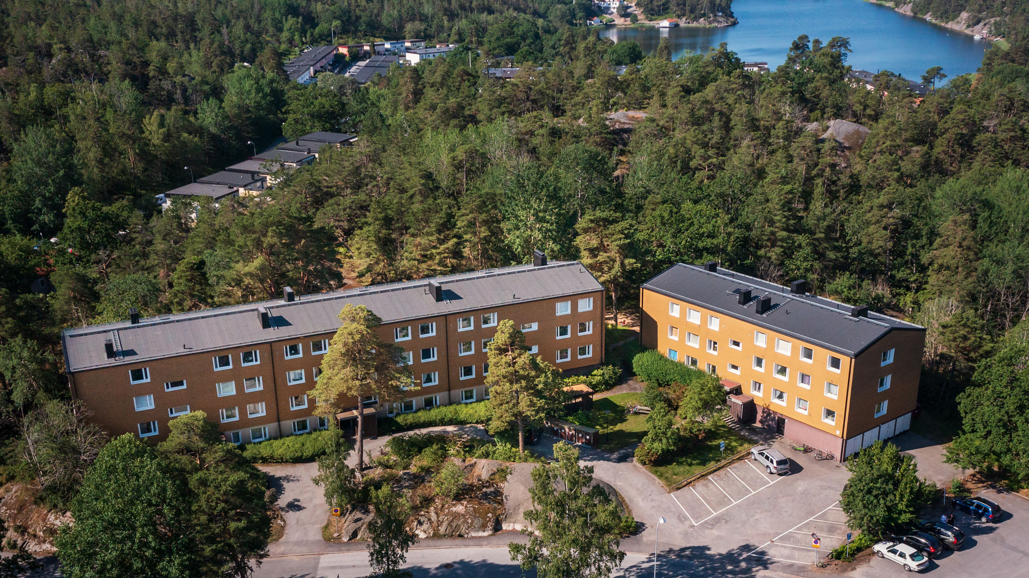 Flygbild över hyreslägenheter på Krokvägen 3 i Nynäshamn, med gula fasader omgivna av skog och vatten. Perfekta hyreslägenheter i en naturskön miljö med närhet till vatten.